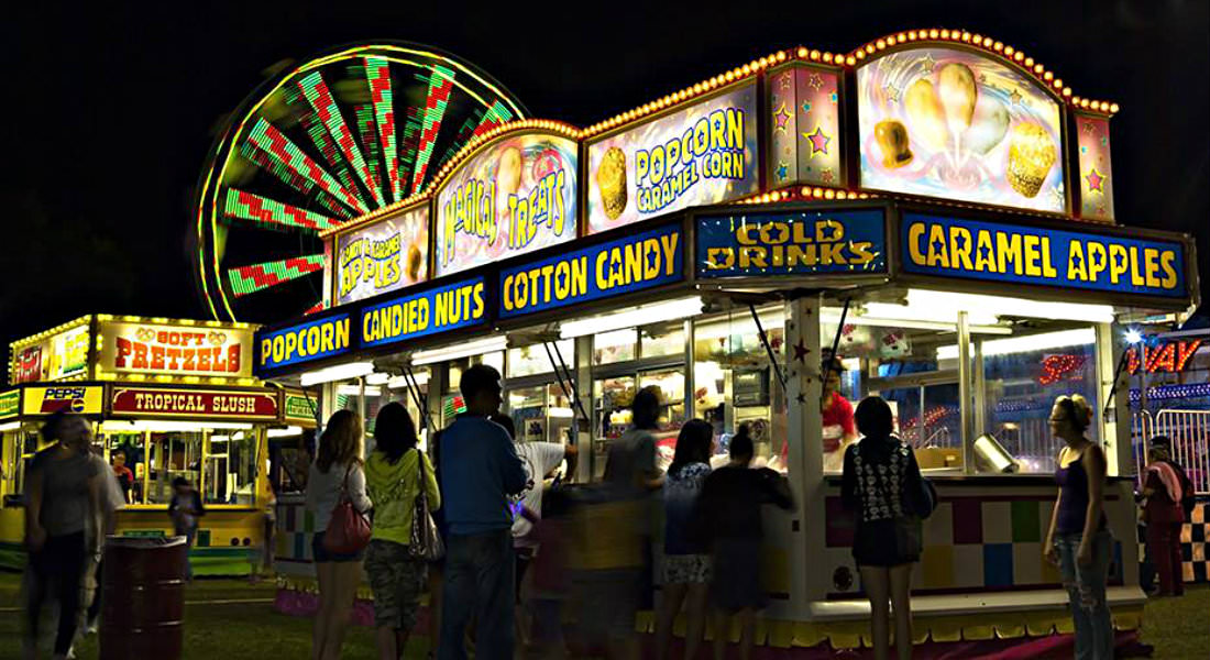 Patrons walking around and gathering around lit concession and carnival rides at night.