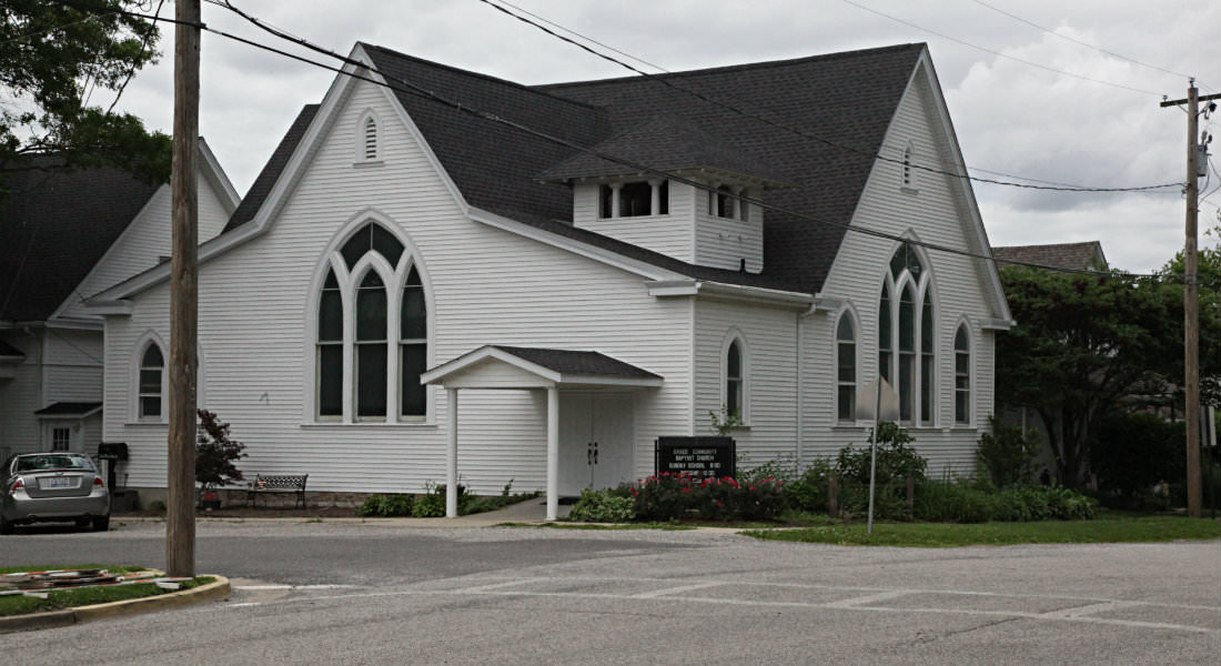 Large white sided church on corner with grey car on angled parking slot on street, sign with services information.