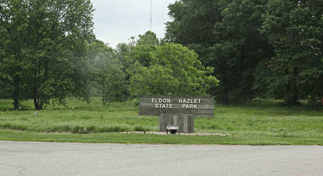 Wood sign along road with white lettering for Eldon Hazlett State Park with a lot of trees behind.