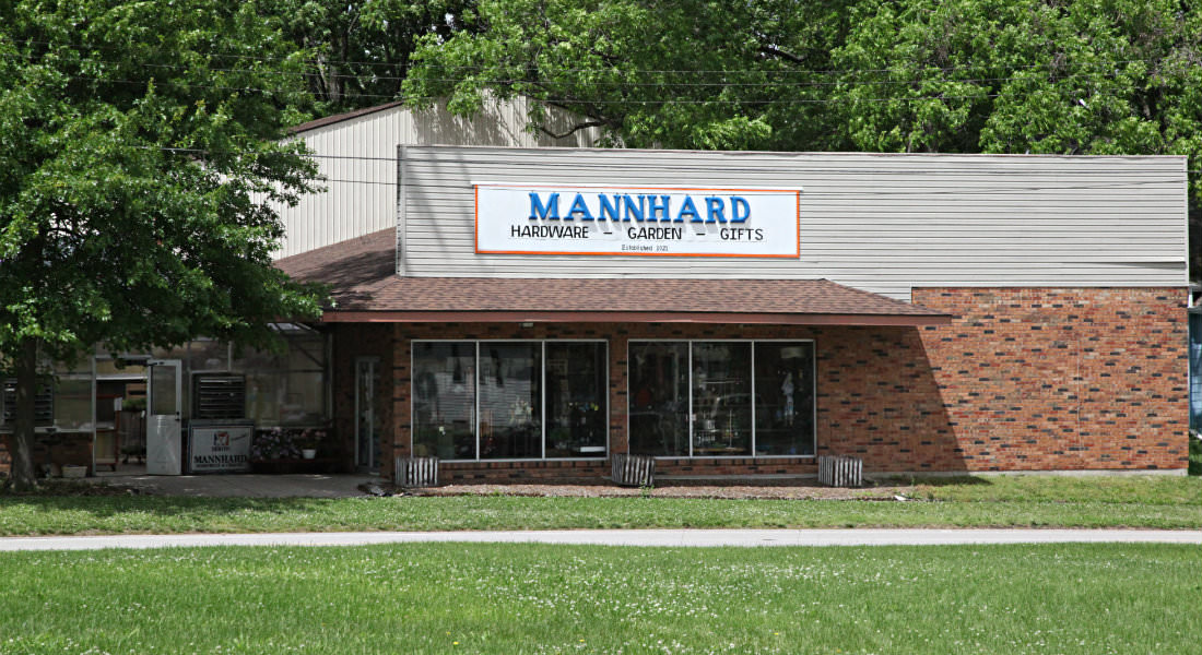 Red brick storefront with tan sided 2nd floor, white sign with blue letters Mannhard Hardware-Garden-Gifts.