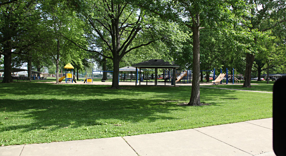 Park with covered shelters and playground equipment under trees, sidewalk surrounding grass.