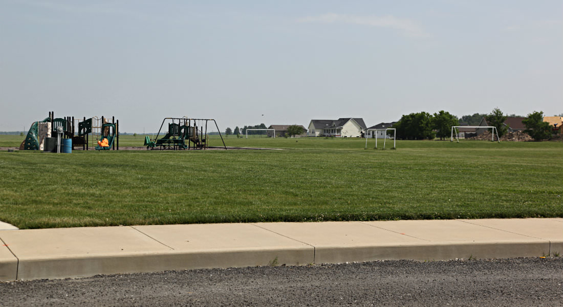 Open grassed area with variety of playground equipment, soccer nets, yellow house in distance.