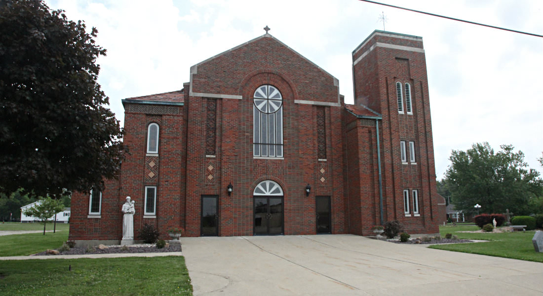 Red brick Church with stained glass windows, cement cross on top, white statue, wide sidewalk in front.