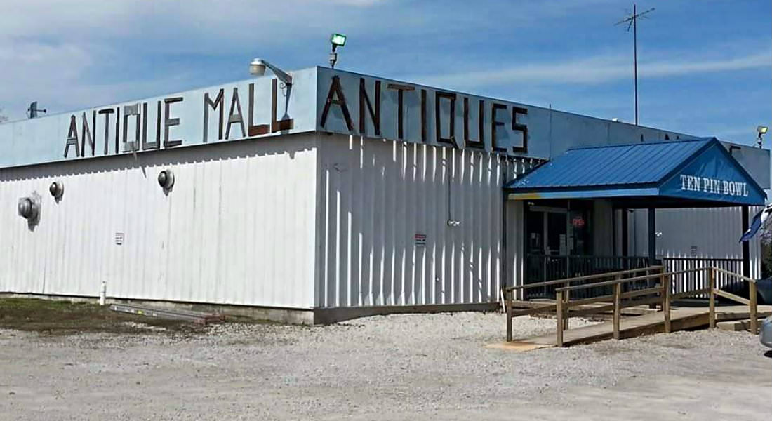 Cream sided building with Antique Mall Antiques with gravel parking area, wood ramp with railings at entrance.