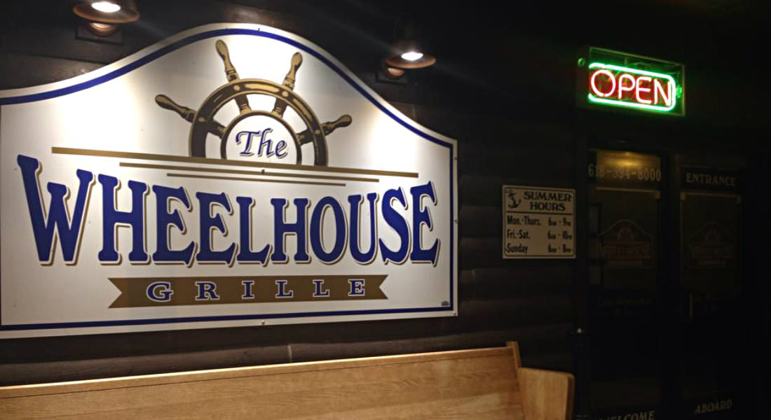 Cream sign with blue lettering The Wheelhouse Grille lit up at night, boat wheel on sign at top.
