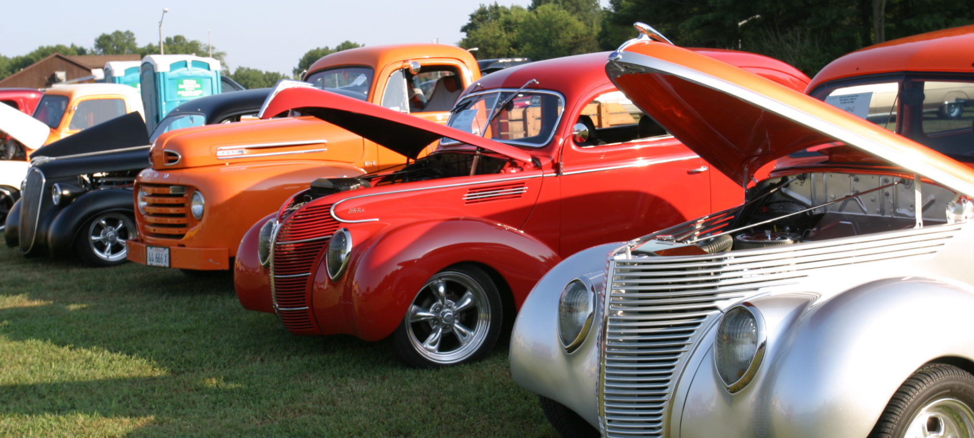 Various colors of classic Antique cars parked on lawn at car show, porta pots, trees.