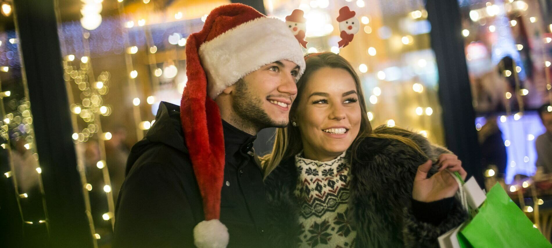 Couple smiling in front of a store’s twinkling lights. Man has Santa hat, and woman has a shopping bag.