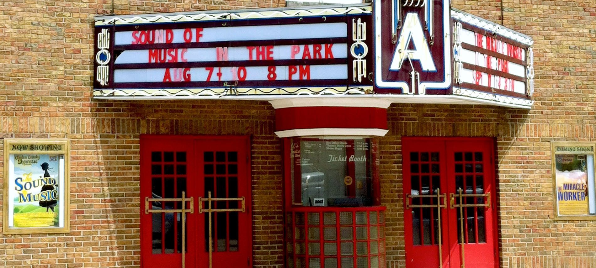 Front of tan brick theatre building with red doors with, ticket booth, billboard announcing Sound of Music In The Park.