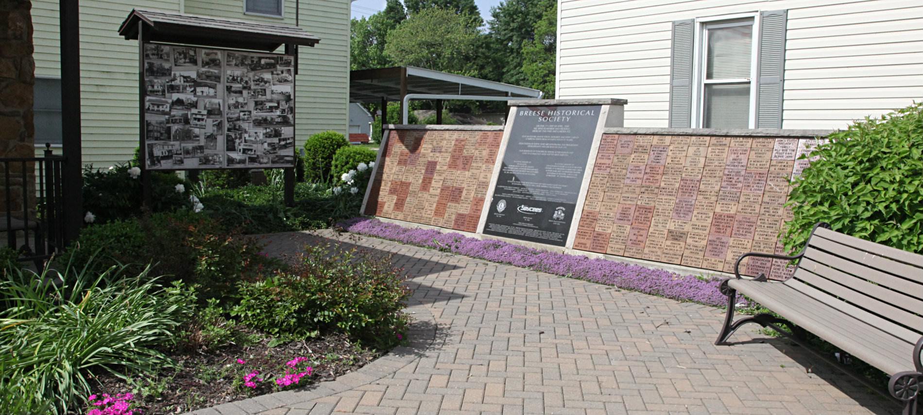 Patterned walkway made out of bricks leading to bulletine board with collage of pictures and on right Breeze Historical Society memorial wall.