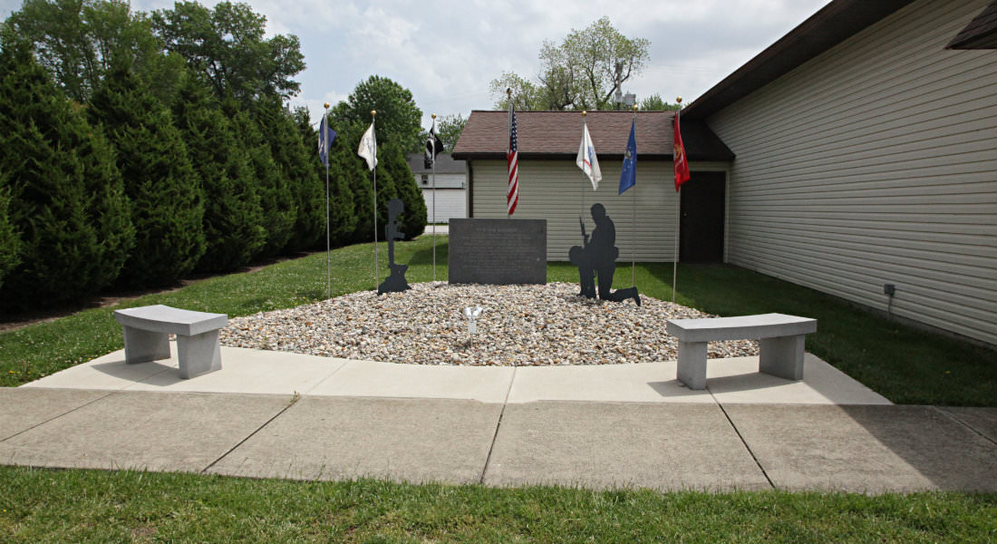 Concrete benches surrounding Memorial Wall with 7 flags on poles, Silhouette figure kneeling in gravel.