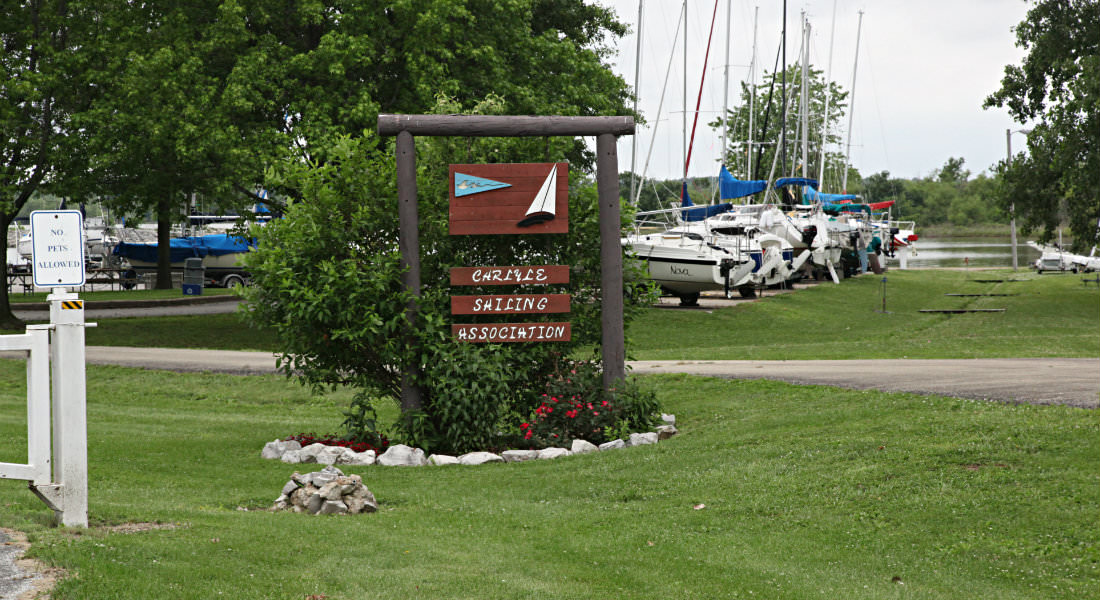 Red wood sign with white letter Carlyle Sailing Association, No Pets Allowed Sign, Sailboats lined up on land.