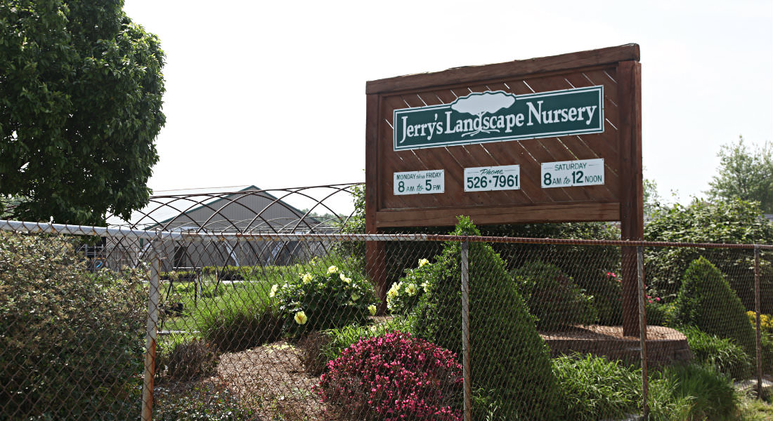 Chain link fence with flowers behind, red wood with green and white sign for Jerry's Landscape Nursery with phone number and hours.
