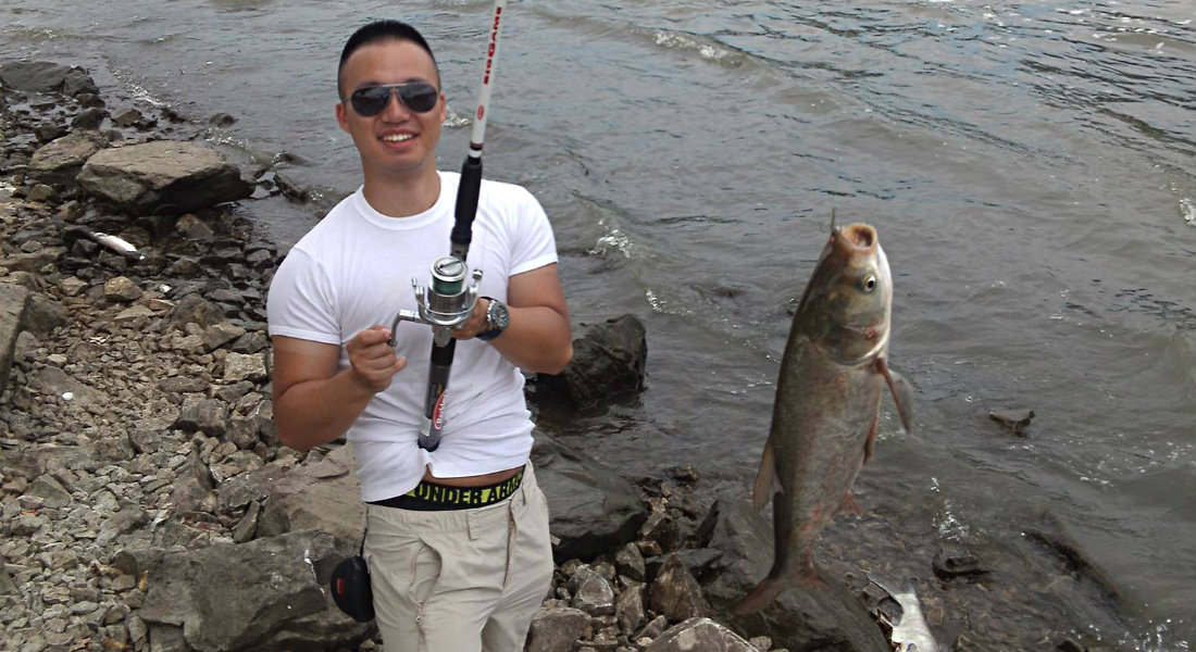 Man wearing white t-shirt and tan shorts on edge of lake holding fishing pole with fish on line.