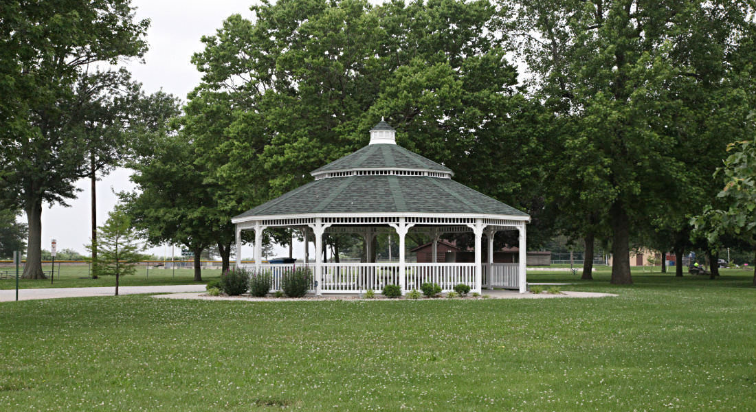 Circular white covered gazebo with white picket fence with tall trees behind, bushes around building.