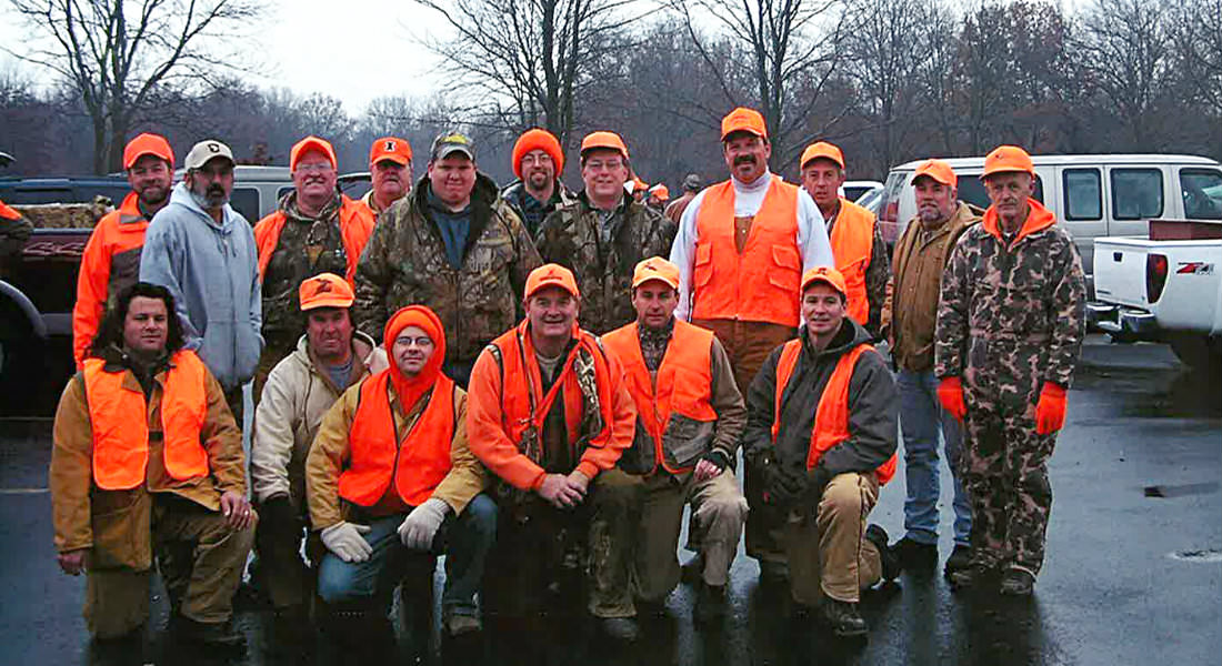 Group of men in coats some wearing camouflage and orange, varias pick-up trucks in parking lot.